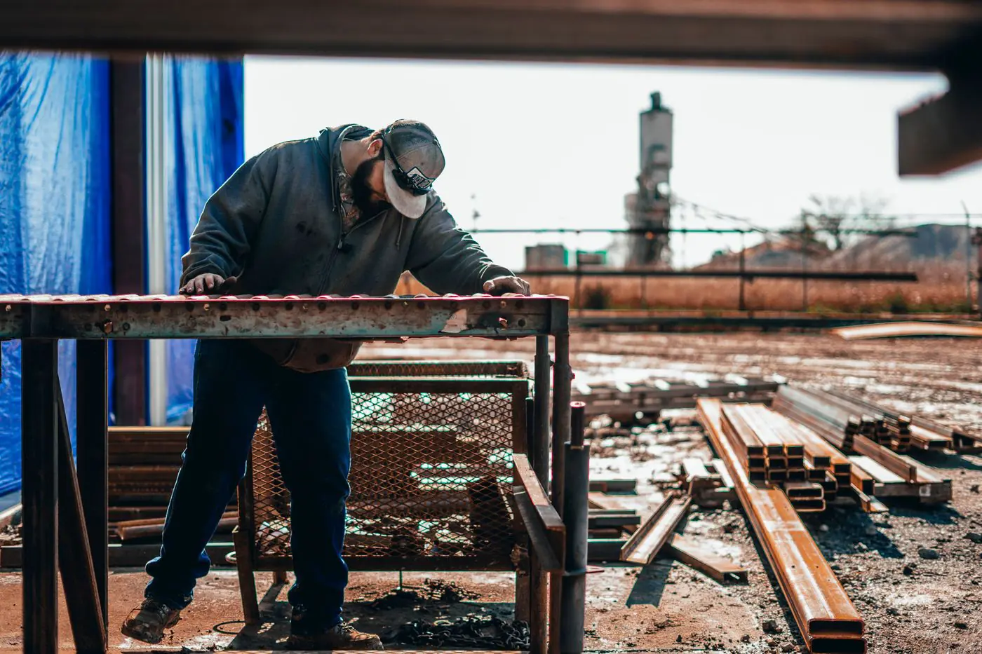 Metal fabricator leaning over a steel frame on trestles in a yard, lengths of square tube and angle stacked alongside