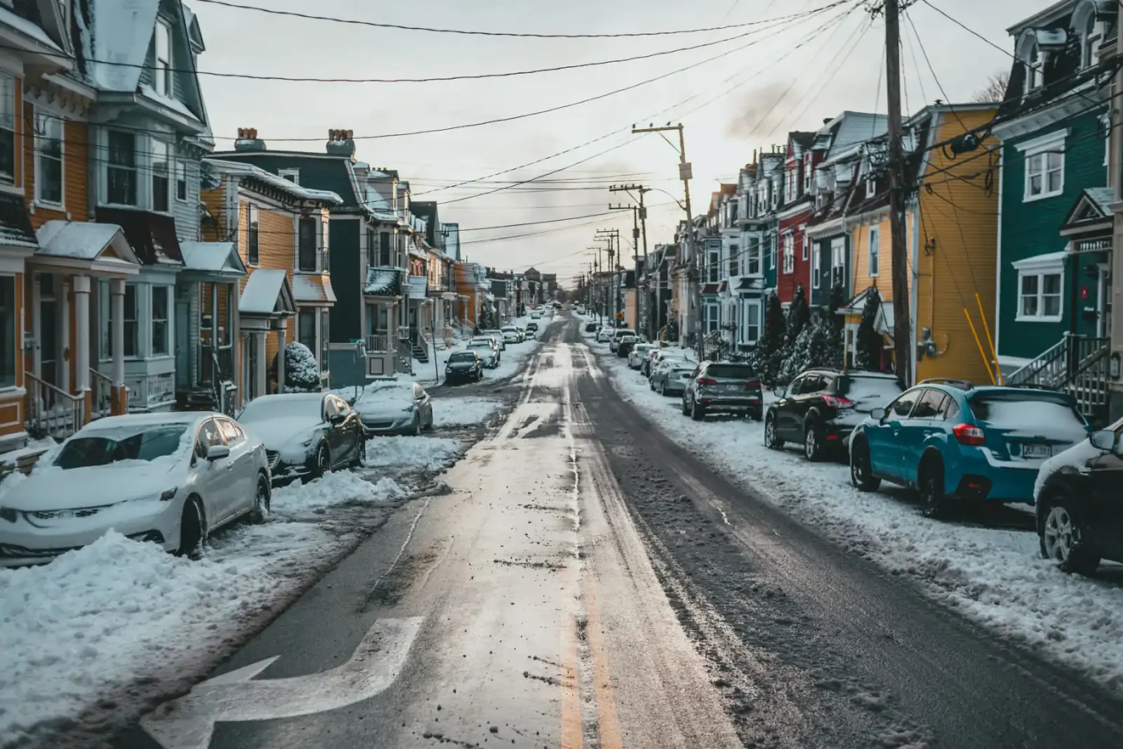Plowed residential street after a snowfall, salt-bleached pavement down the middle and snow berms banked over the parked cars at the curb