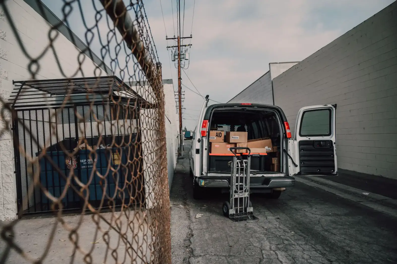 Service van loaded with parts parked in an alley beside a chain-link commercial gate and enclosure