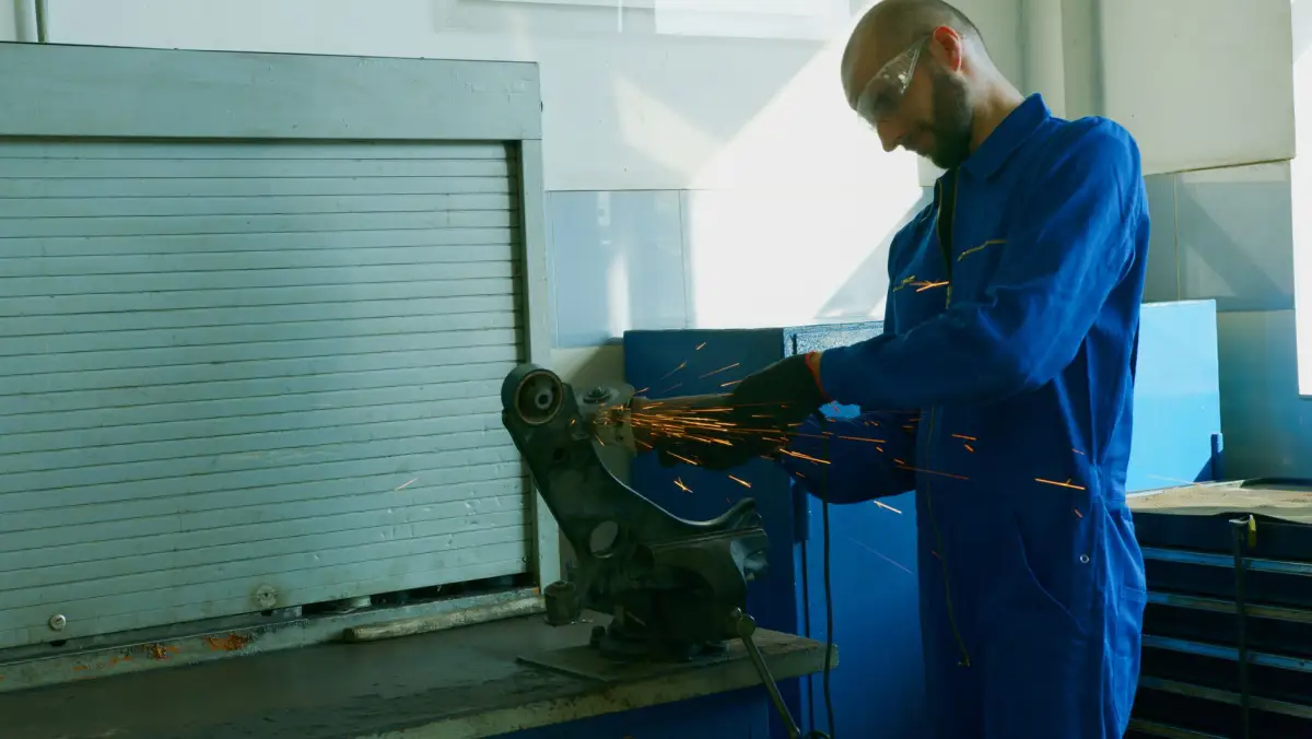 Technician in coveralls and safety glasses grinding a steel component on a bench grinder