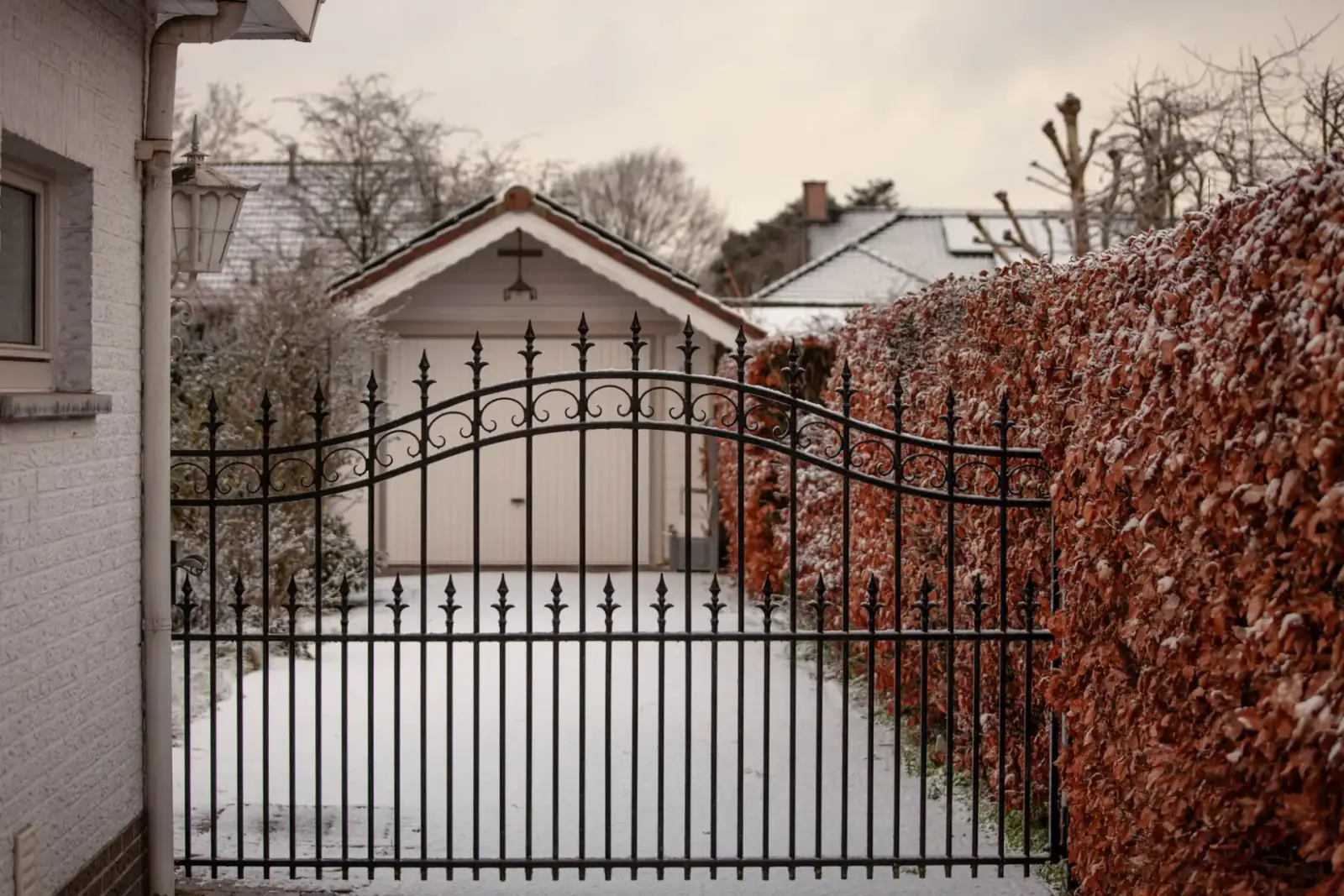 Black spear-top double swing gate with an arched top rail closing a driveway between a garage and a copper beech hedge