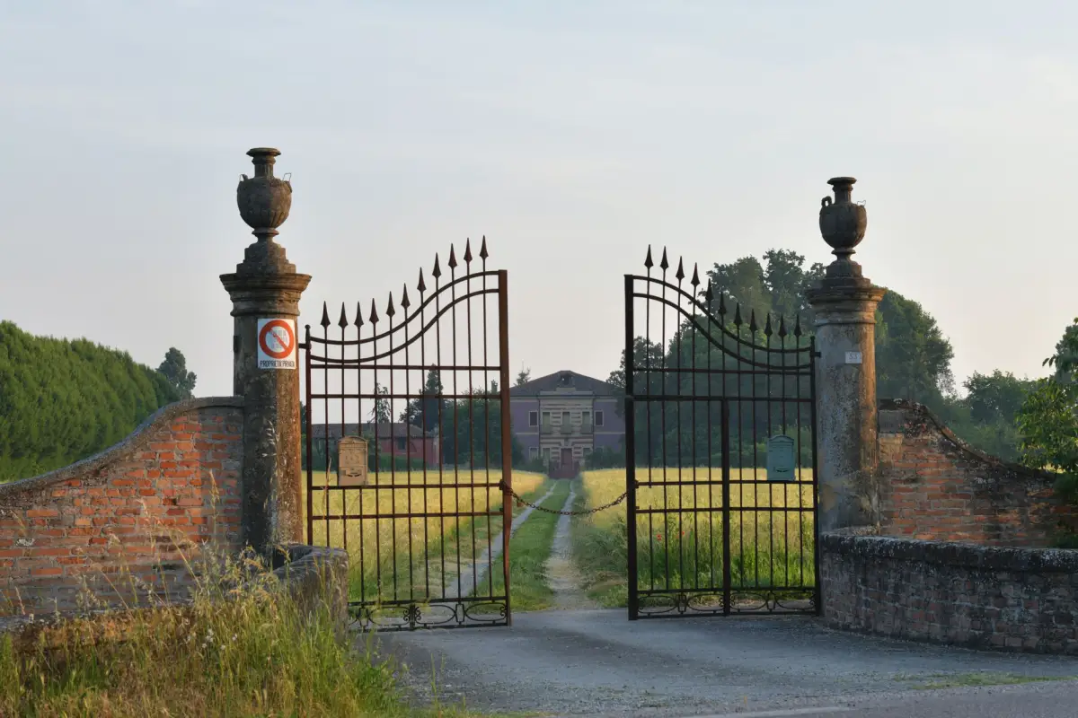 Double iron driveway gate standing open between brick piers with urn finials at the head of a gravel drive