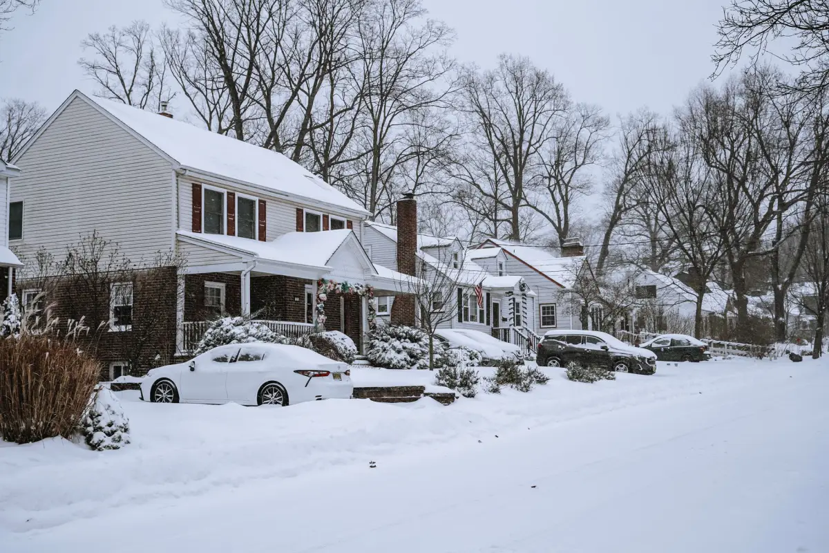 Snow-covered residential street with buried cars and plow berms across the driveway entrances