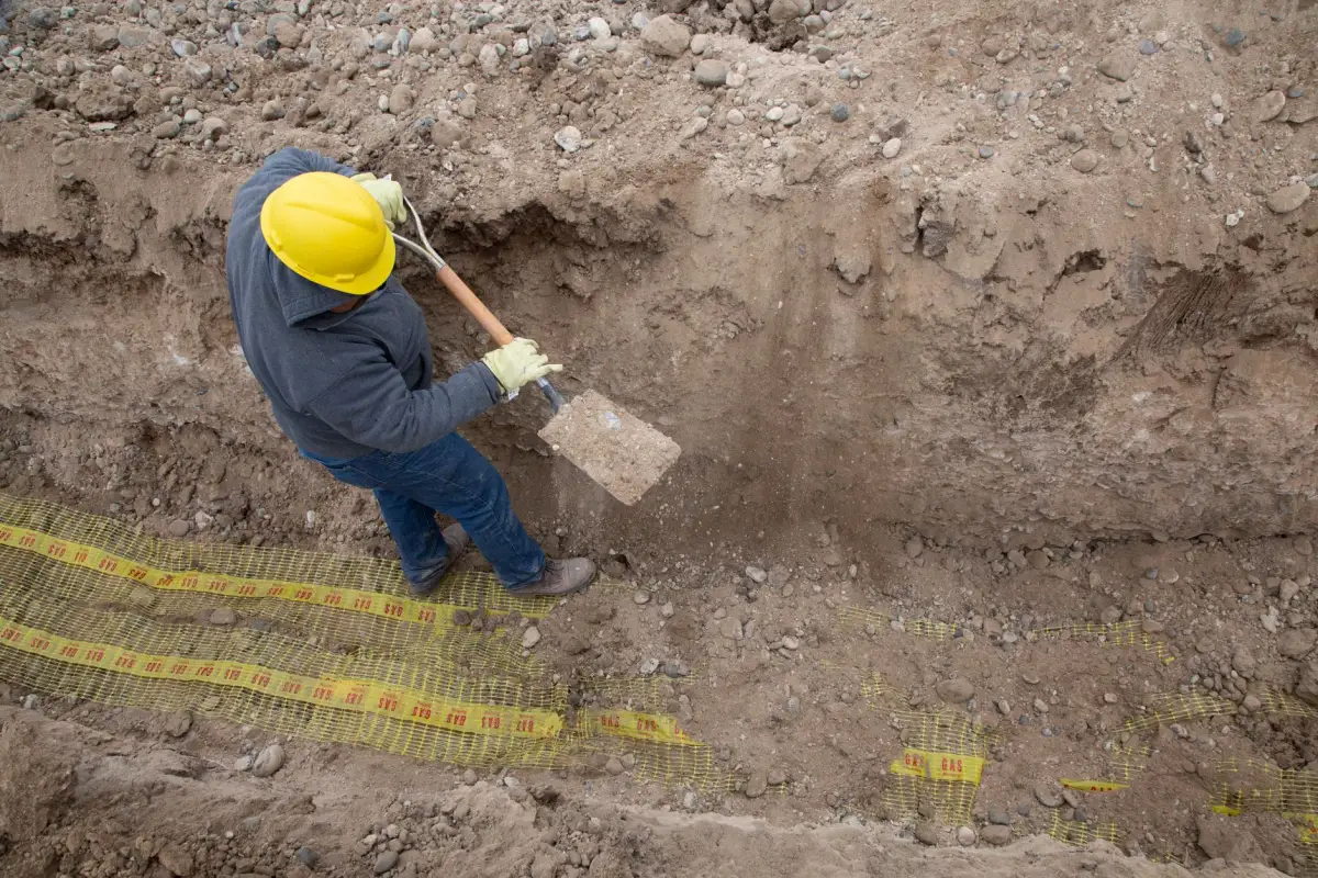 Worker in a hard hat digging a conduit trench with warning tape laid along the excavation