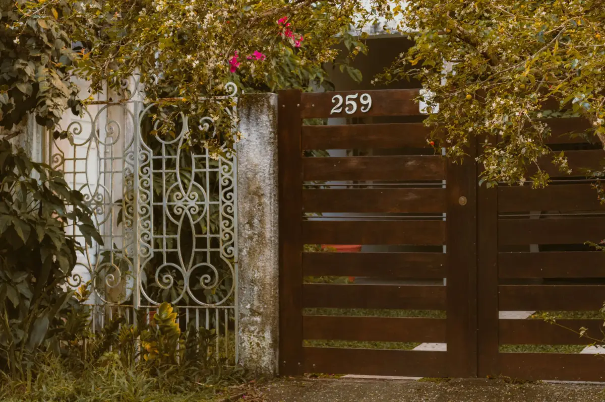 Modern horizontal-slat metal entry gate with an ornamental screen panel and house number beside a planted wall
