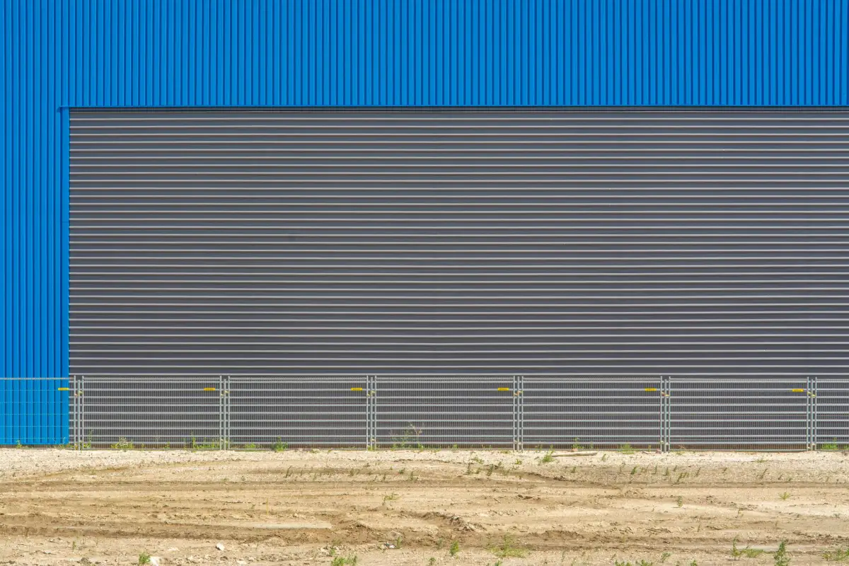 Long metal perimeter fence and slide gate run alongside a blue-clad warehouse building