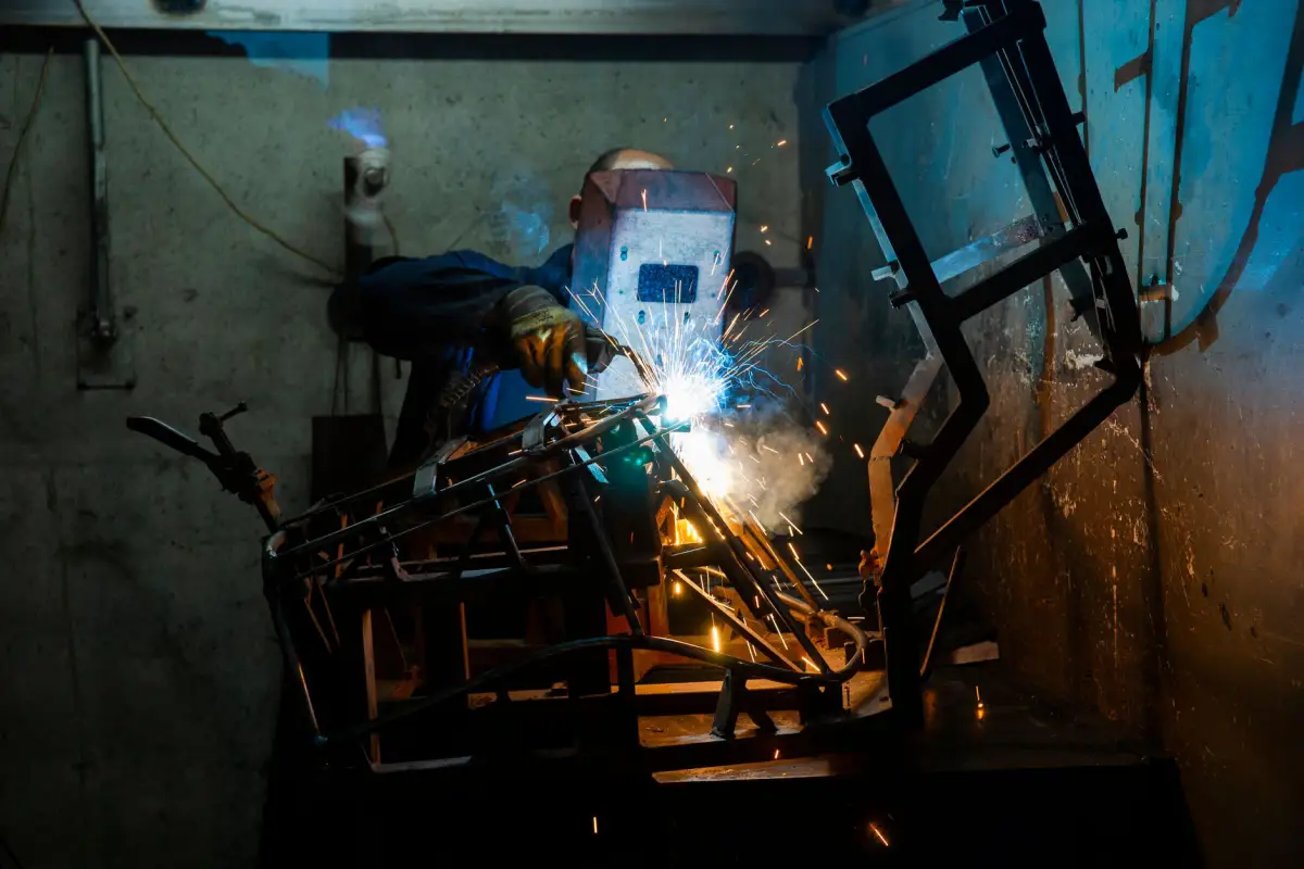 Fabricator in a welding hood welding a rectangular steel swing gate frame in a workshop