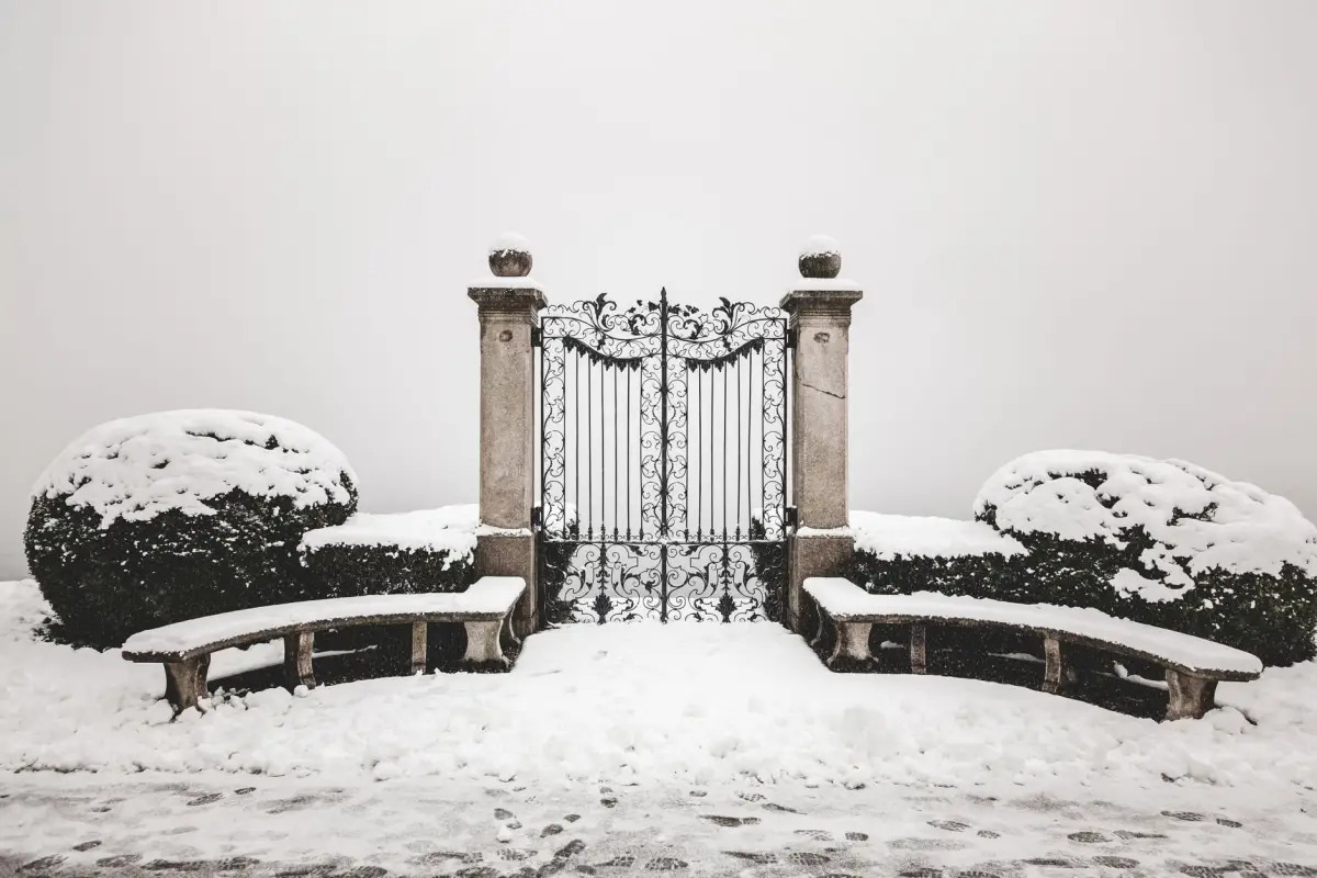 Ornate wrought iron gate between stone piers with deep snow banked against the hedges either side